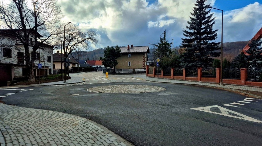 Rondo z kostki granitowej na skrzyżowaniu, otoczone asfaltową jezdnią. W tle zabudowa jednorodzinna i zieleń. Widoczne znaki drogowe i oświetlenie uliczne.