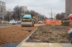 Walec drogowy ubija żwirowe podłoże pod chodnik. W tle widoczne budynki miejskie i zaparkowane samochody. Prace drogowe w miejskiej przestrzeni.