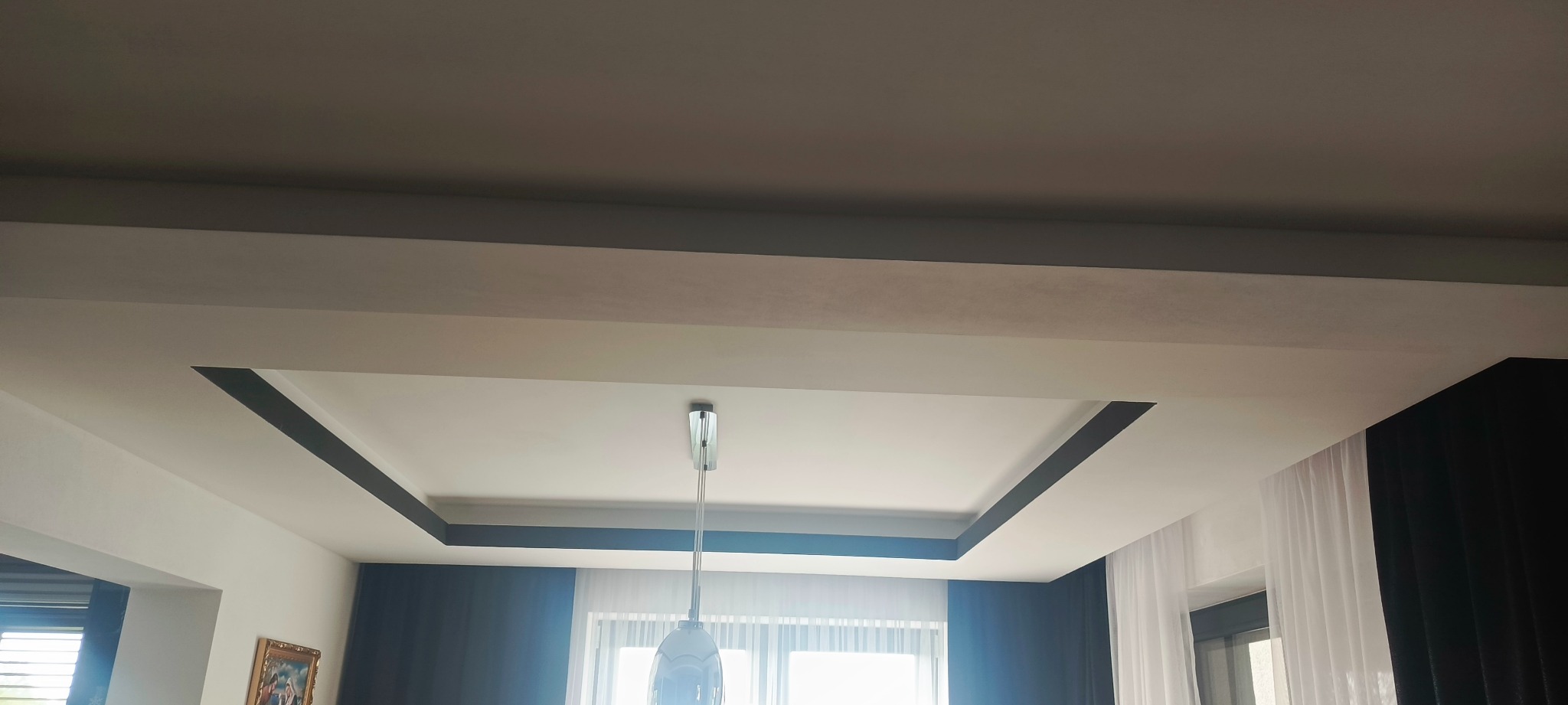 Biały sufit z czarną, geometryczną dekoracją i lampą wiszącą. Widok z dołu, z fragmentami okien i zasłon w tle. Nowoczesny design wnętrza.