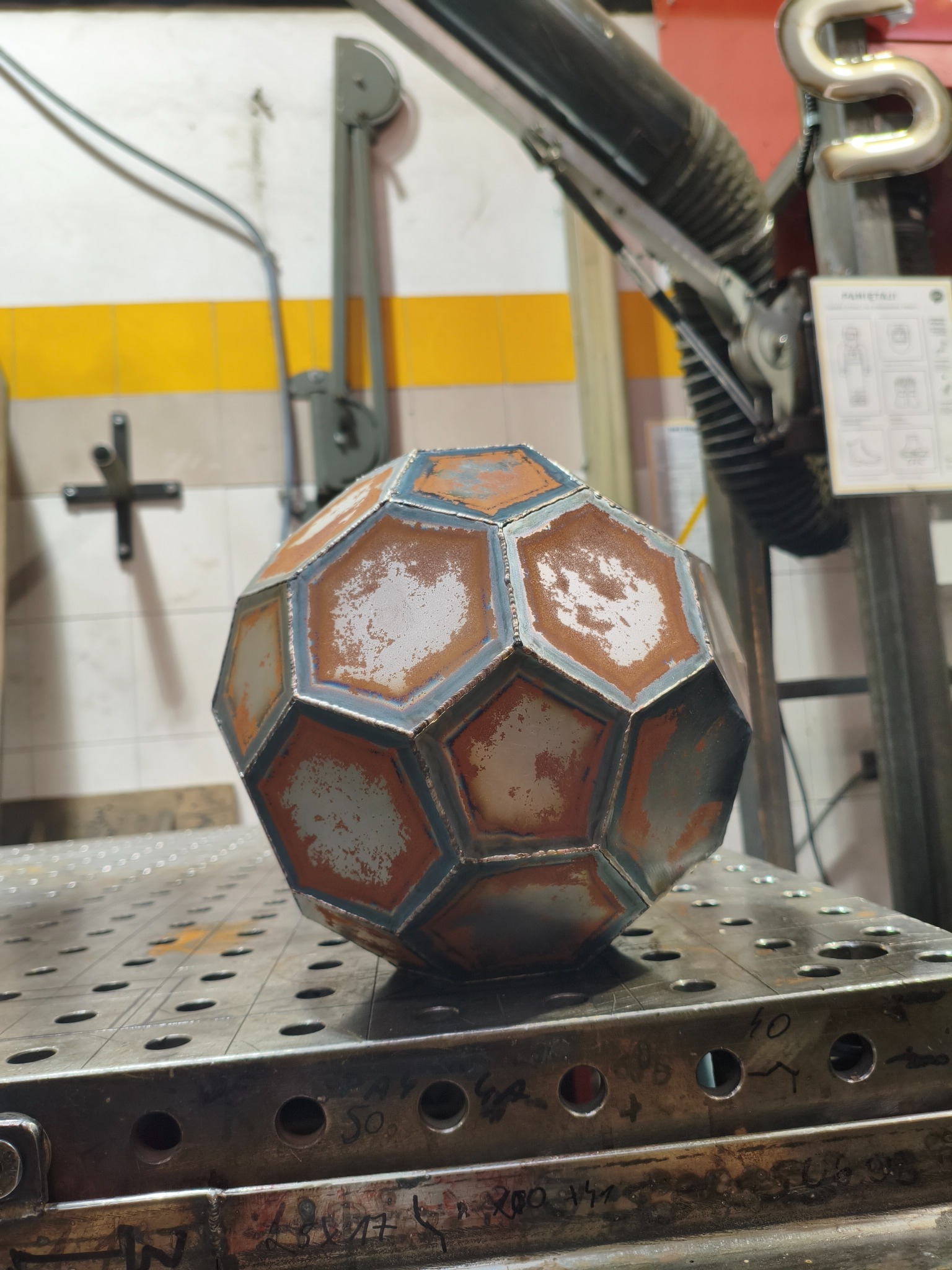 Spawana metalowa kula z wielokątów na perforowanym stole spawalniczym. Widoczne ślady spawania i rdzy, co sugeruje proces obróbki metalu. W tle warsztat.