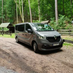 Szary bus Renault Trafic z polską rejestracją G1 TUTTI, zaparkowany na leśnym parkingu obok drewnianej wiaty i płotu, w otoczeniu drzew.
