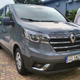 TuttiCars Paweł Juchniewicz - Szary Renault Trafic z polską tablicą rejestracyjną na brukowanym podjeździe, w tle przyczepa kempingowa i budynek z klimatyzacją.