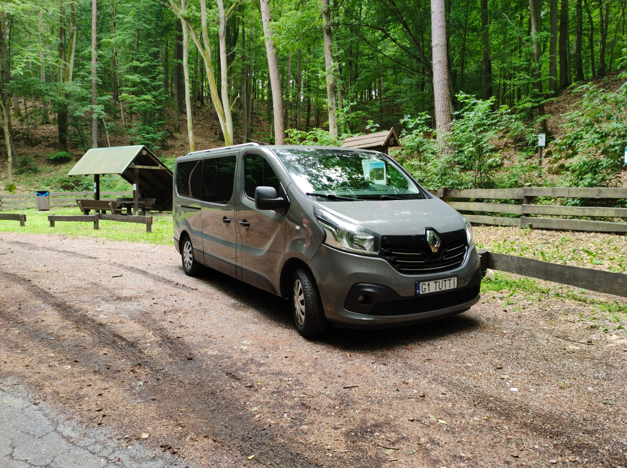 Szary bus Renault Trafic z polską rejestracją G1 TUTTI, zaparkowany na leśnym parkingu obok drewnianej wiaty i płotu, w otoczeniu drzew.