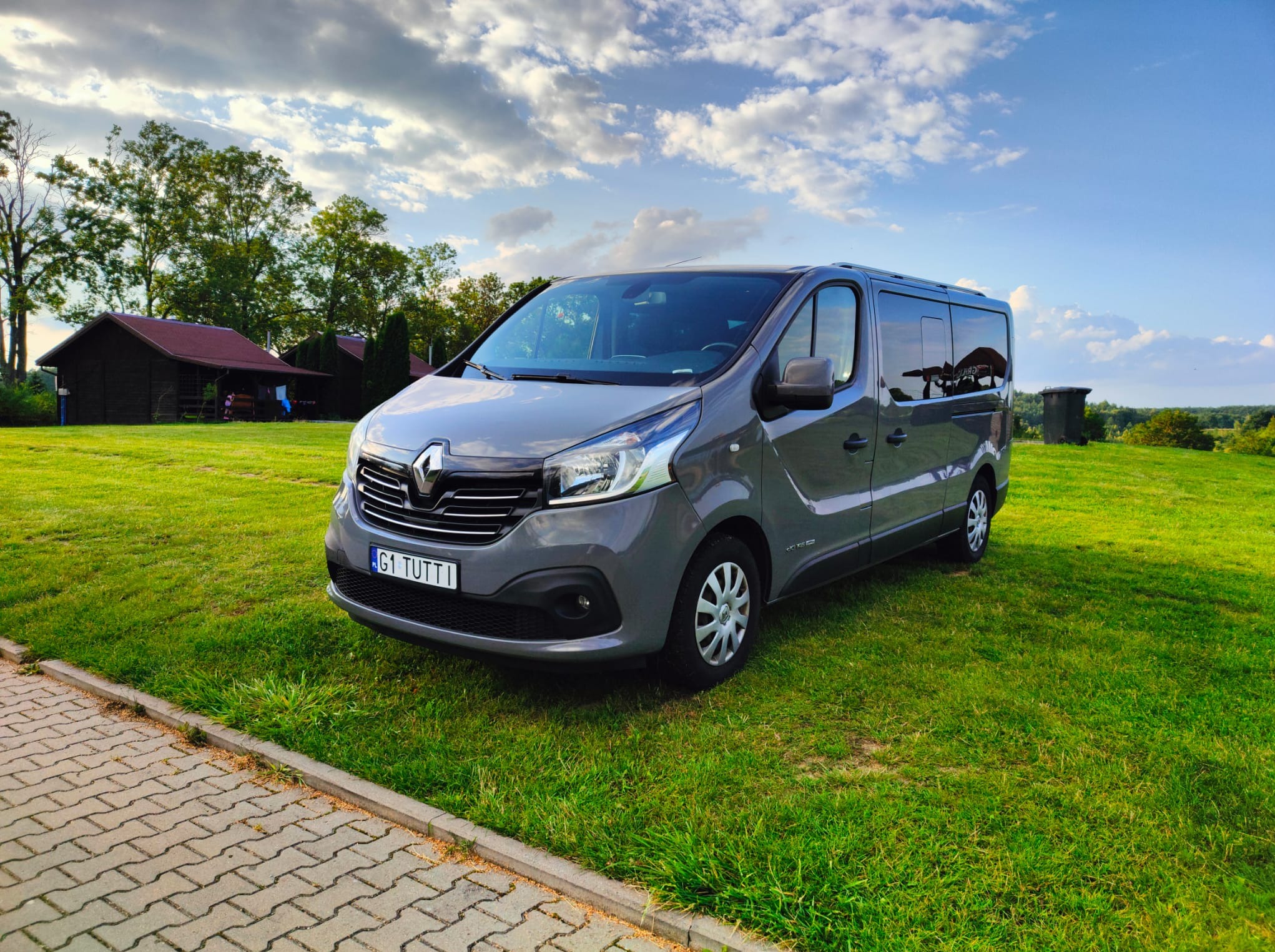 Szary Renault Trafic na zielonej trawie, błękitne niebo z chmurami w tle. Tablica rejestracyjna 'G1 TUTTI'.