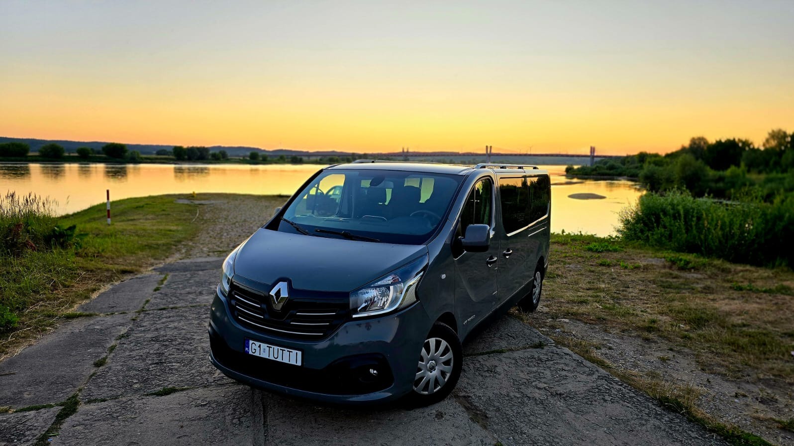 Szary bus Renault na tle rzeki i zachodzącego słońca. Widoczny most w oddali i zielona roślinność. Tablica rejestracyjna G1 TUTTI.