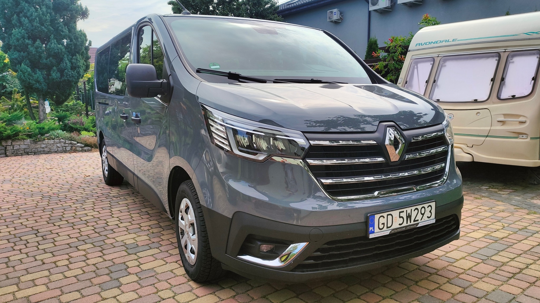 Szary Renault Trafic z polską tablicą rejestracyjną na brukowanym podjeździe, w tle przyczepa kempingowa i budynek z klimatyzacją.