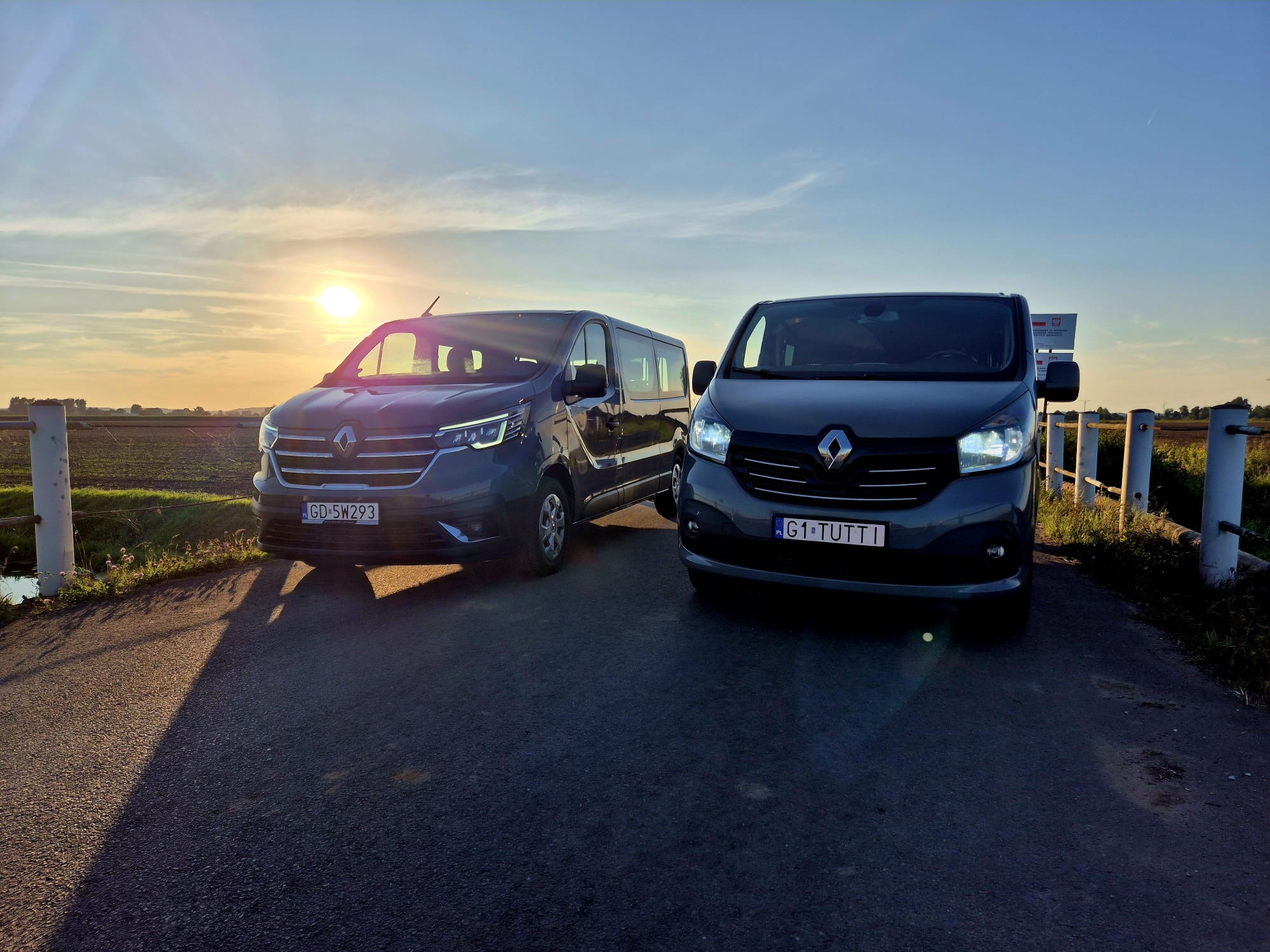 Dwa busy Renault na tle zachodzącego słońca, z widocznymi tablicami rejestracyjnymi i polskimi oznaczeniami. Pojazdy zaparkowane na drodze, w tle pola.
