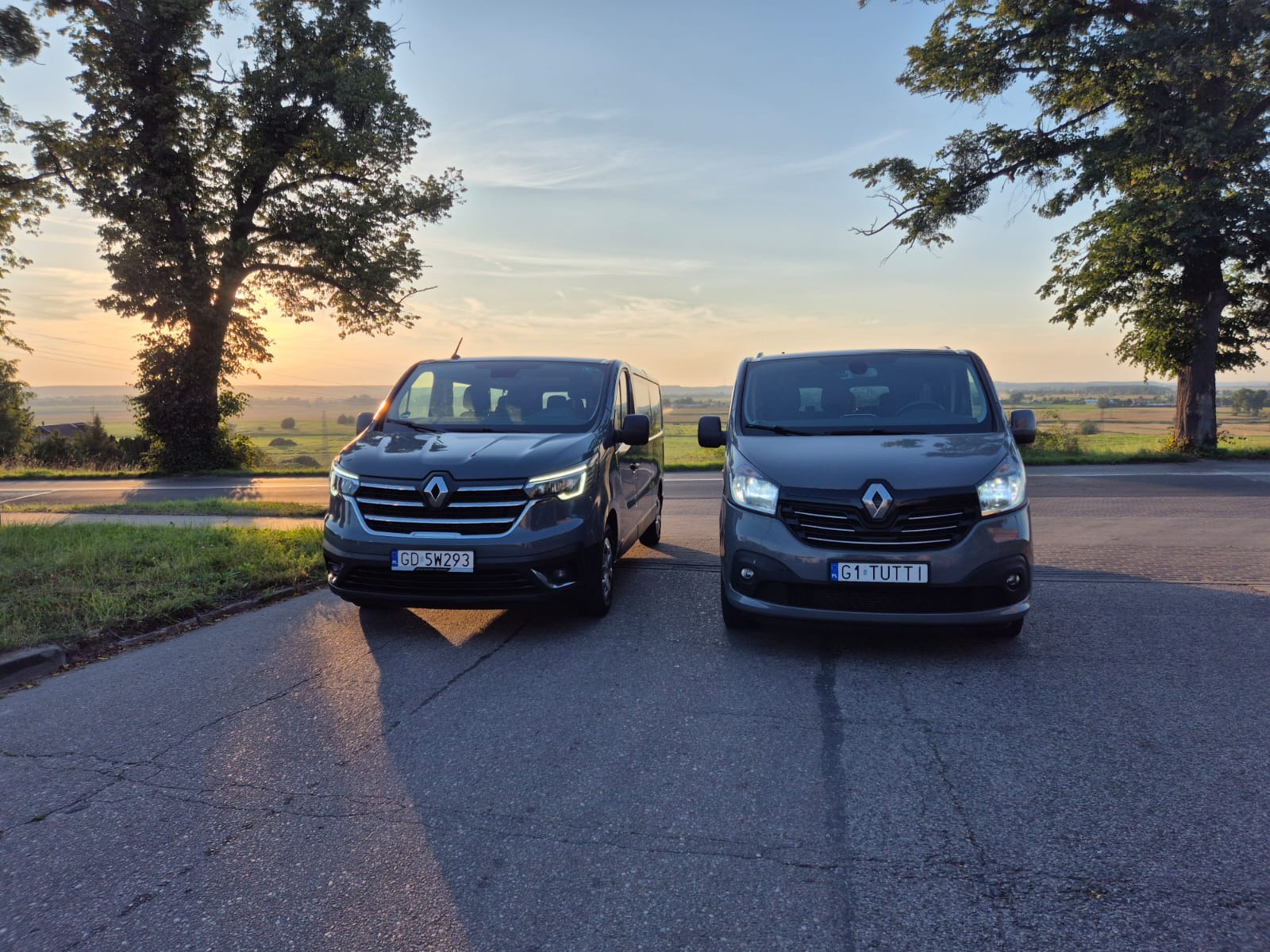 Dwa szare busy Renault na tle malowniczego krajobrazu o wschodzie słońca. Pierwszy plan zajmuje asfaltowa droga z widocznymi pęknięciami i śladami użytkowania.