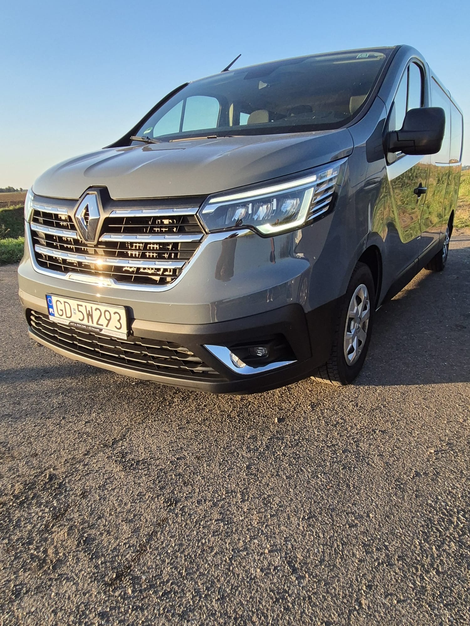 Szary bus Renault Trafic z polską tablicą rejestracyjną GD 5W293, zaparkowany na żwirowej drodze w słoneczny dzień. Widok z przodu.