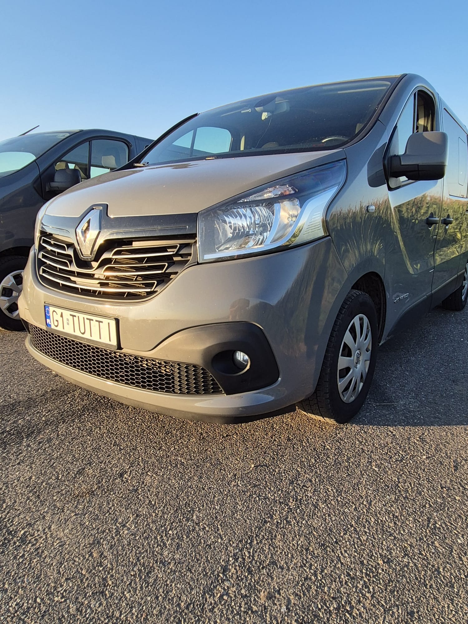Szary Renault Trafic z włoską tablicą rejestracyjną G1-TUTTI, zaparkowany na zewnątrz w słoneczny dzień. Widoczny fragment drugiego pojazdu.