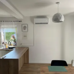 Biały klimatyzator na ścianie nad stołem w jasnej kuchni. Widoczny geometryczny żyrandol i dekoracje ścienne. Nowoczesny design.