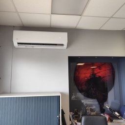 Spokój W Zasięgu Ręki Bartłomiej Olejniczak - Biały klimatyzator AUX zamontowany na szarej ścianie biura, widoczny fragment sufitu kasetonowego, biurko z krzesłem i szafka z żaluzjami.