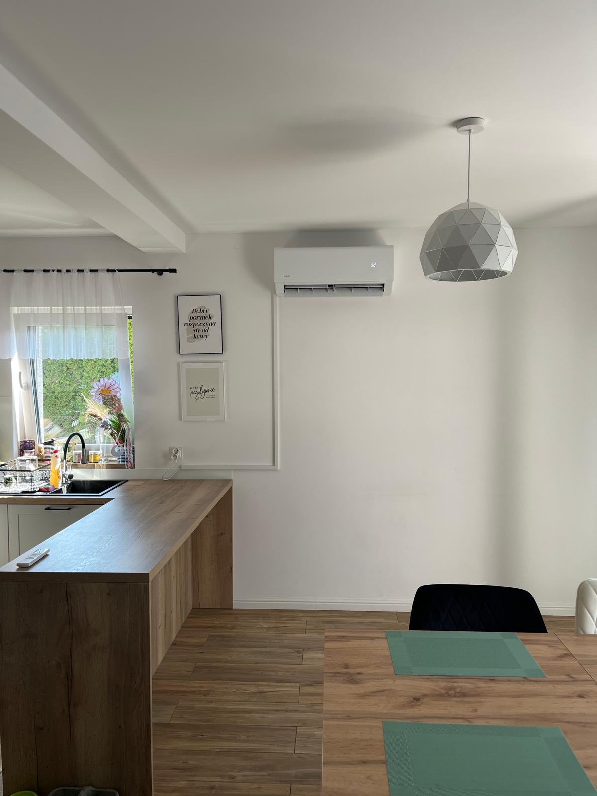 Biały klimatyzator na ścianie nad stołem w jasnej kuchni. Widoczny geometryczny żyrandol i dekoracje ścienne. Nowoczesny design.
