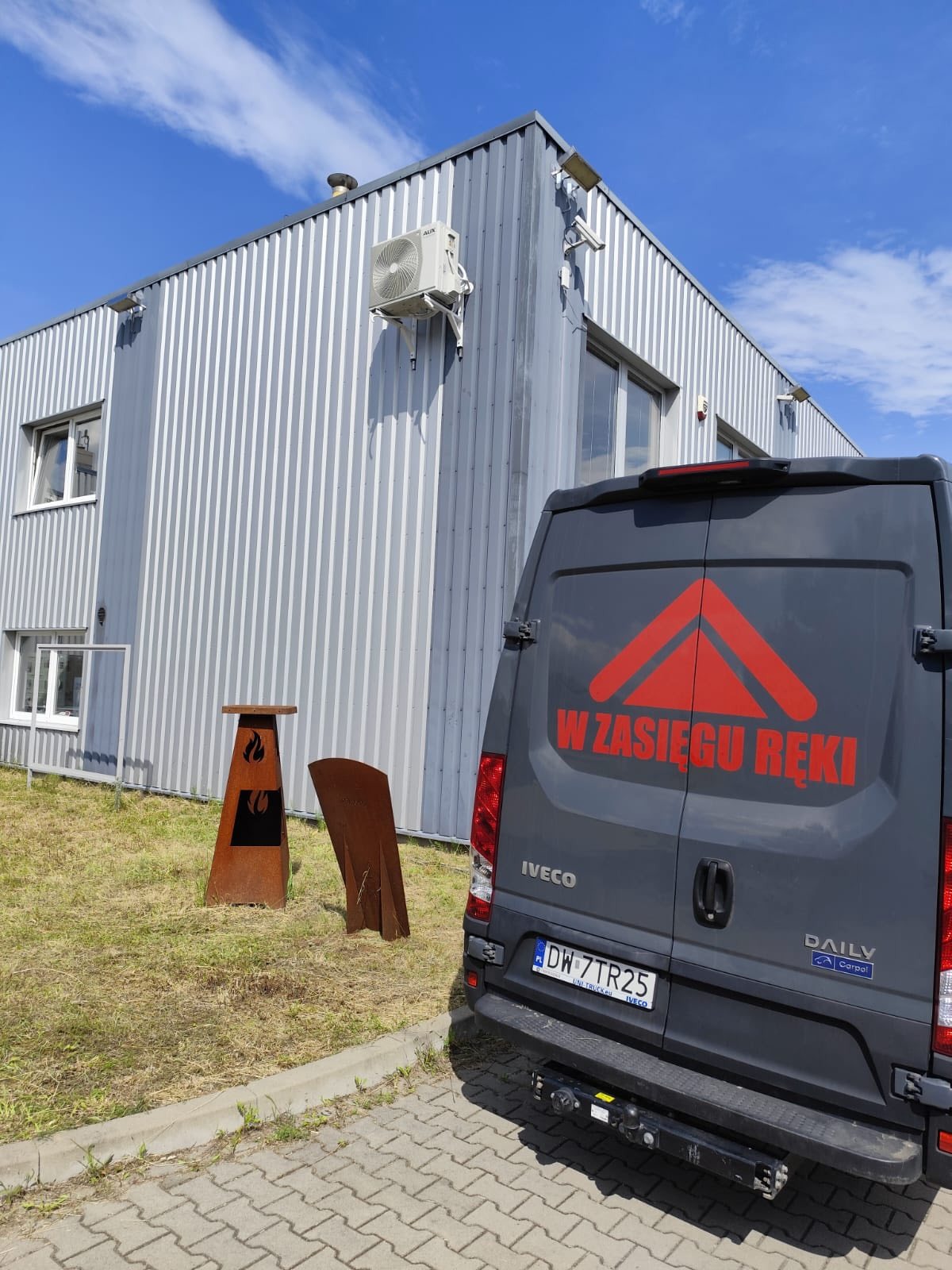 Szara hala z klimatyzatorem AUX na ścianie, obok zaparkowany szary bus Iveco z czerwonym logo i napisem. Na trawniku dekoracyjne, metalowe elementy.