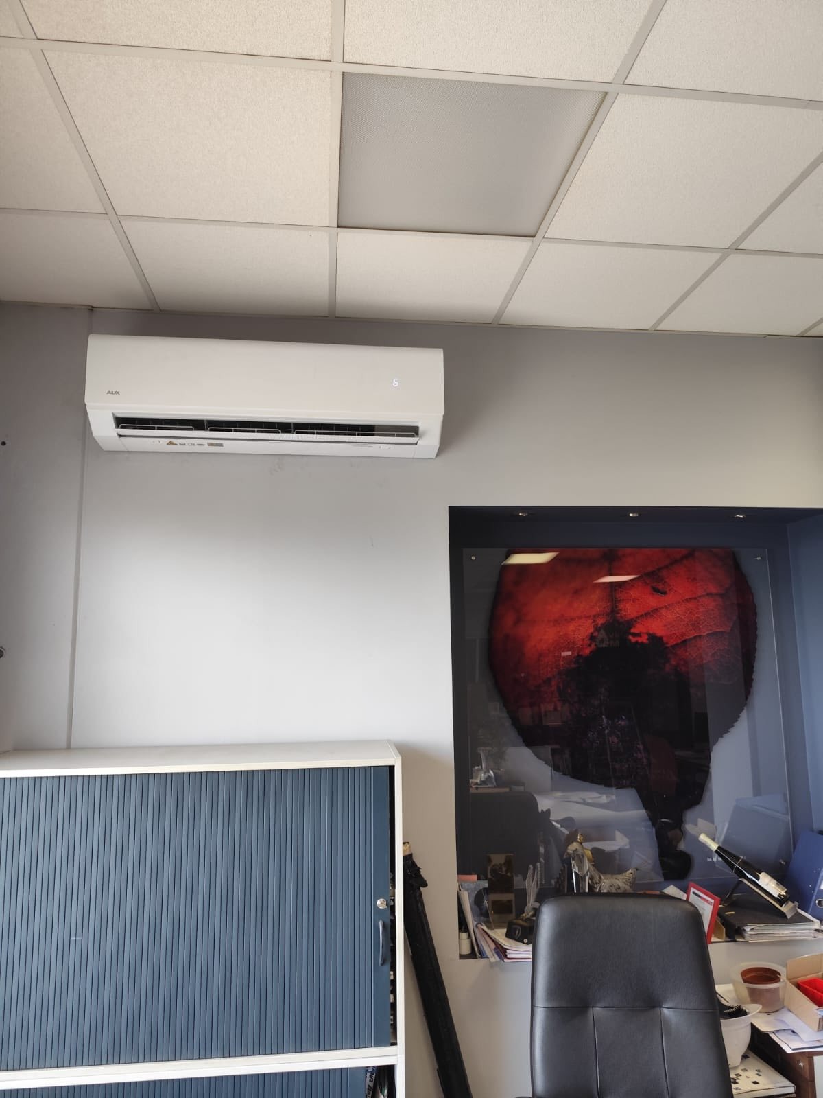 Biały klimatyzator AUX zamontowany na szarej ścianie biura, widoczny fragment sufitu kasetonowego, biurko z krzesłem i szafka z żaluzjami.