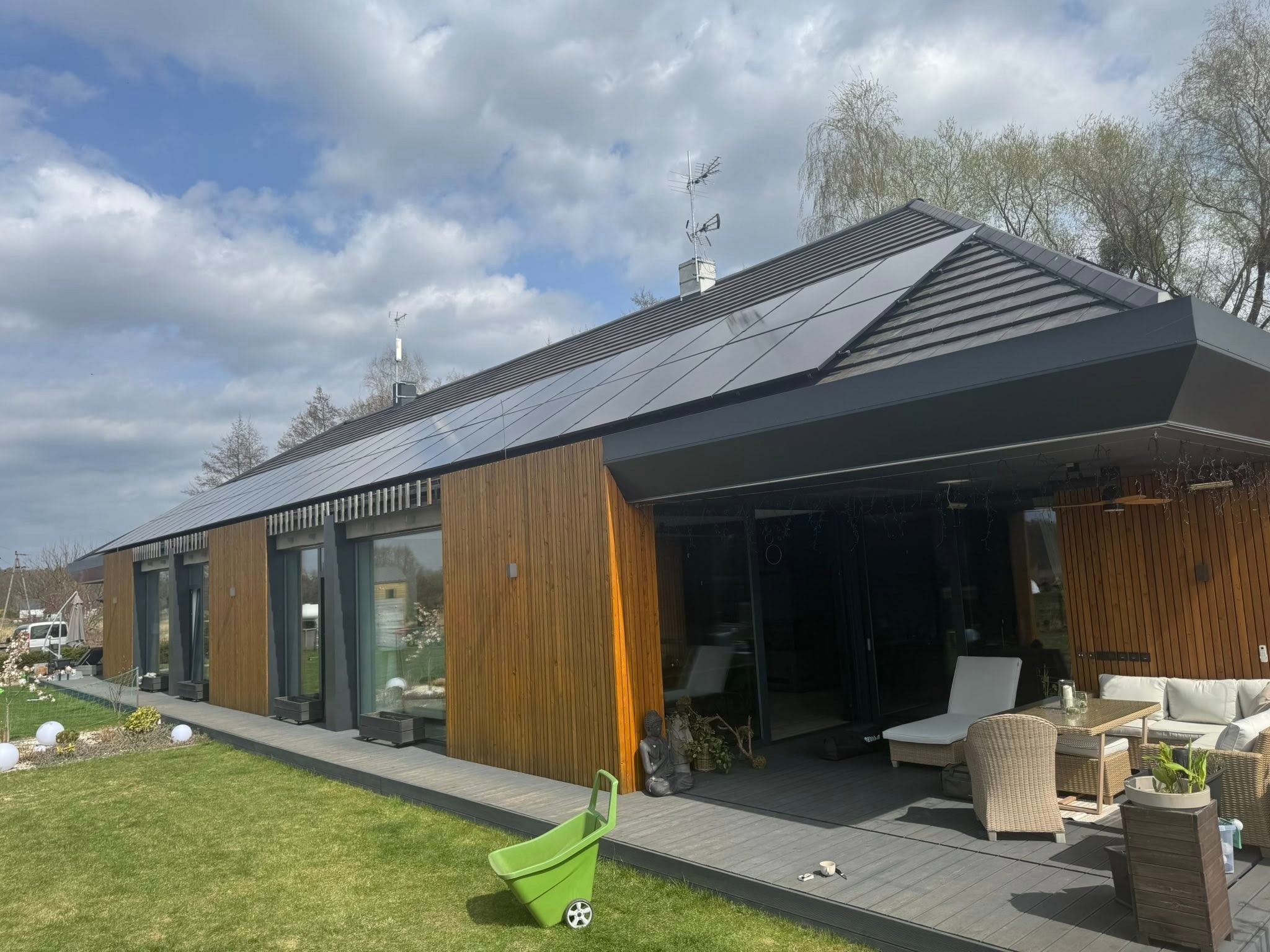 Nowoczesny dom jednorodzinny z panelami fotowoltaicznymi na dachu, widok z zewnątrz, drewniana elewacja, taras z meblami ogrodowymi, wózek ogrodowy na trawniku.