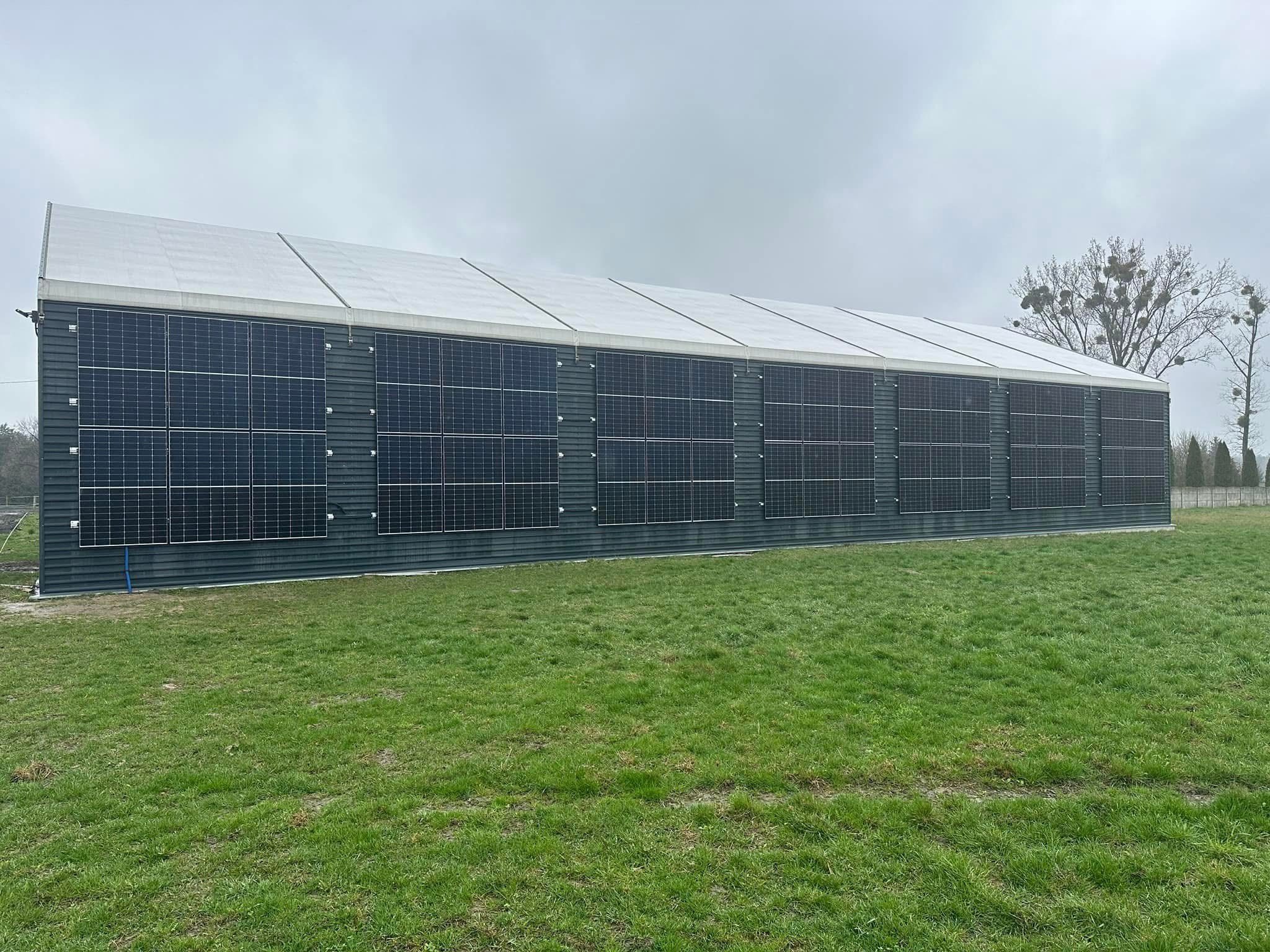 Duży budynek gospodarczy z panelami fotowoltaicznymi na ścianach, otoczony zielonym trawnikiem w pochmurny dzień. Widoczne drzewa w tle.