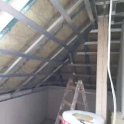 Skośne poddasze w trakcie ocieplania wełną mineralną, widoczny stelaż metalowy, drabina i rolka materiału izolacyjnego z częściowo odpakowaną folią.