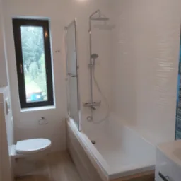 Jasna łazienka po renowacji z wanną z prysznicem, toaletą wiszącą i oknem z widokiem na zieleń, oświetlona pojedynczą żarówką.