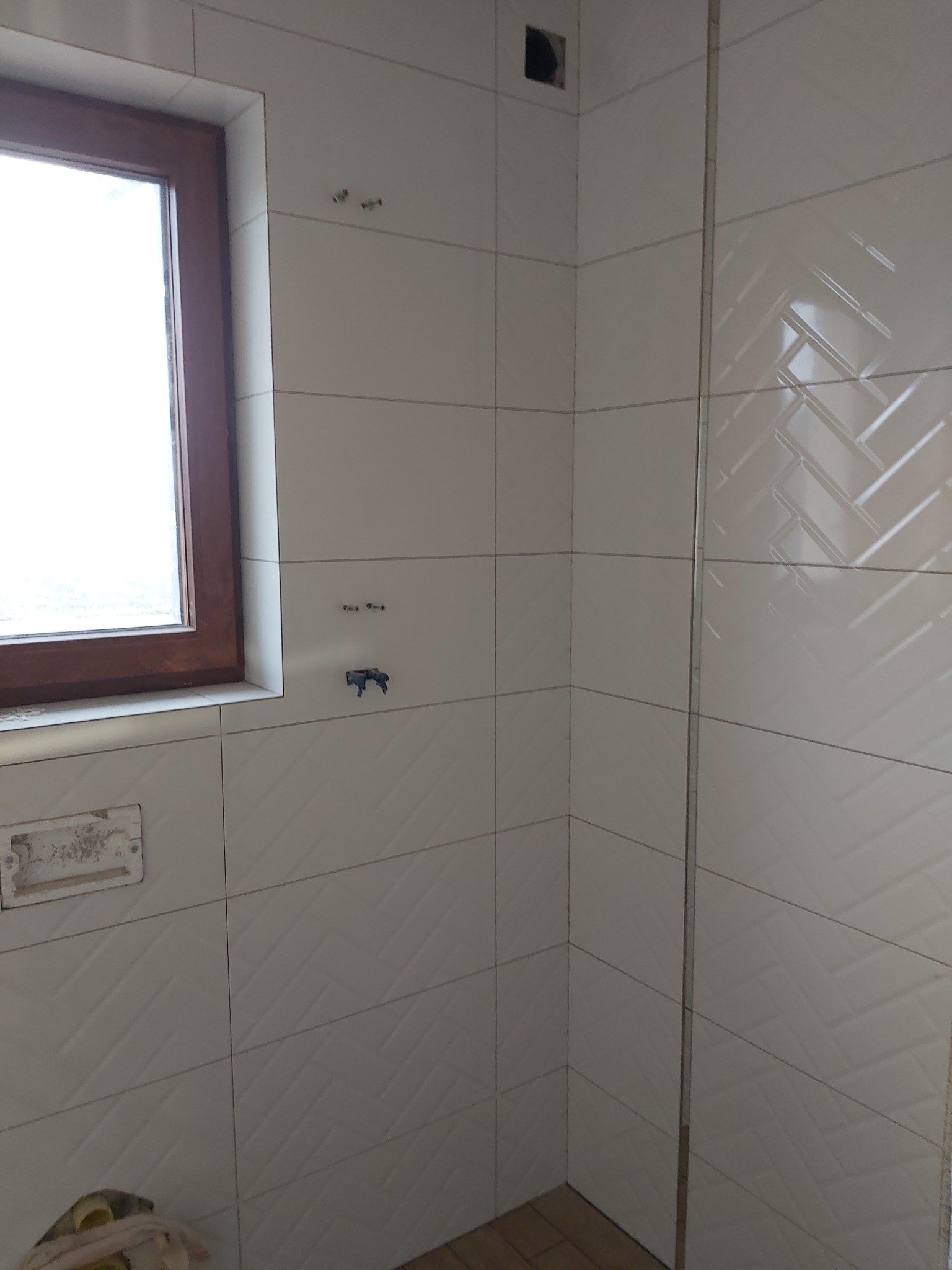 Ściany łazienki wyłożone białymi, prostokątnymi płytkami z subtelnym wzorem jodełki, widoczne przygotowane punkty instalacyjne i fragment drewnianego okna.