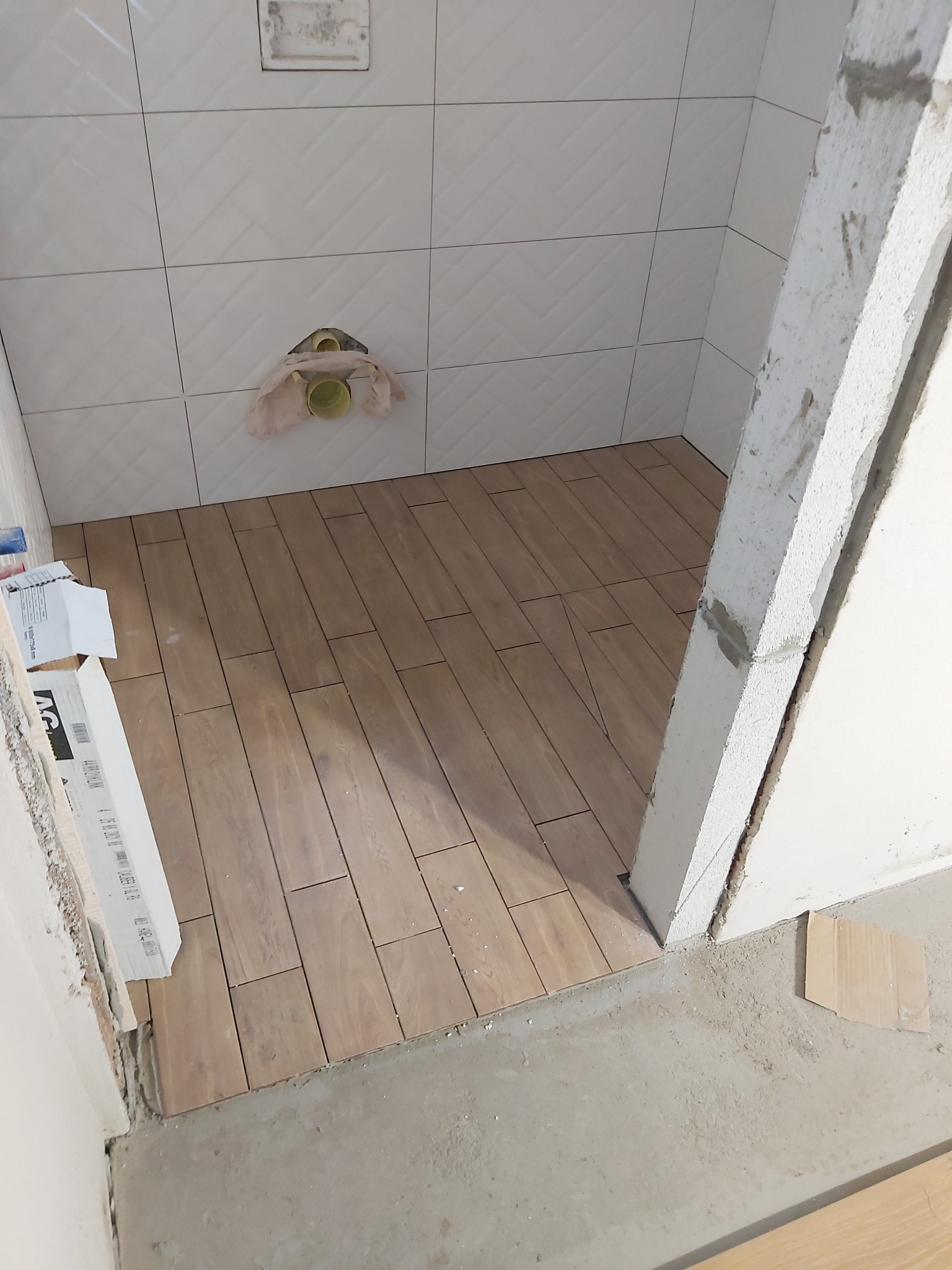 Ułożona podłoga z płytek imitujących drewno w trakcie prac w łazience, widoczne białe płytki na ścianach z przygotowanym miejscem na instalację sanitarną.