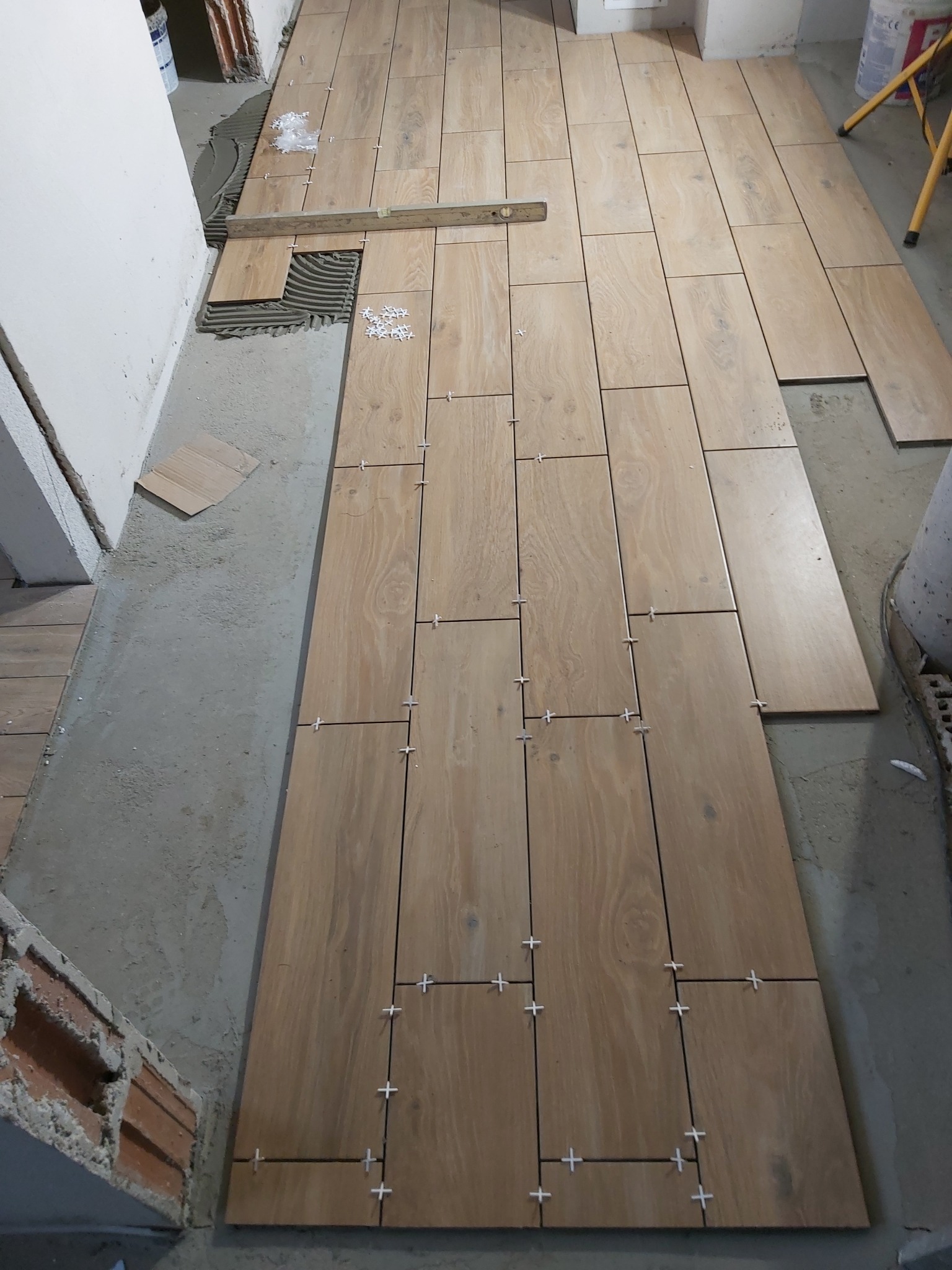 Układanie podłogi z płytek imitujących drewno, widoczne krzyżyki dystansowe, poziomica i resztki zaprawy.