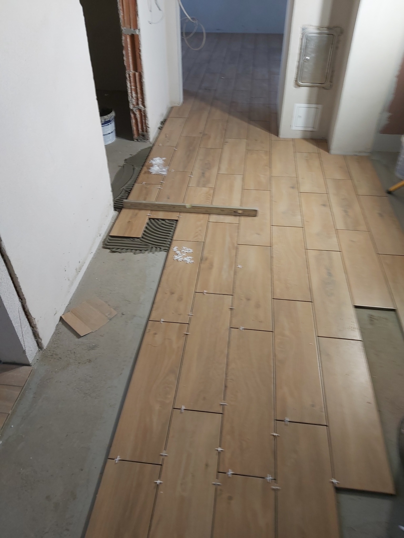 Układanie podłogi z drewnopodobnych płytek w korytarzu, widoczne krzyżyki dystansowe, poziomica i odsłonięty fragment podłoża z naniesionym klejem.