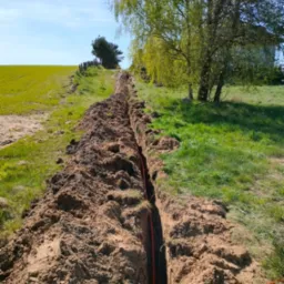 Wykopany rów w ziemi, prawdopodobnie pod instalacje, z widocznymi pomarańczowymi kablami. W tle zielone pole i drzewa w słoneczny dzień.