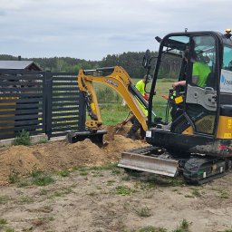 SYSTEM-MEDIA SPÓŁKA Z OGRANICZONĄ ODPOWIEDZIALNOŚCIĄ - Żółto-czarna mini koparka Carter podczas wykopu pod instalacje przy ogrodzeniu posesji. Widoczny operator w kabinie i pracownik z łopatą. Praca w toku.