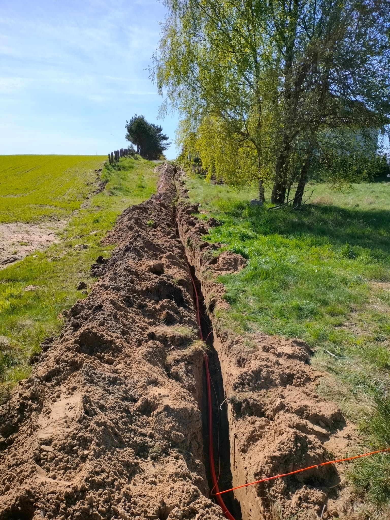 Wykopany rów w ziemi, prawdopodobnie pod instalacje, z widocznymi pomarańczowymi kablami. W tle zielone pole i drzewa w słoneczny dzień.