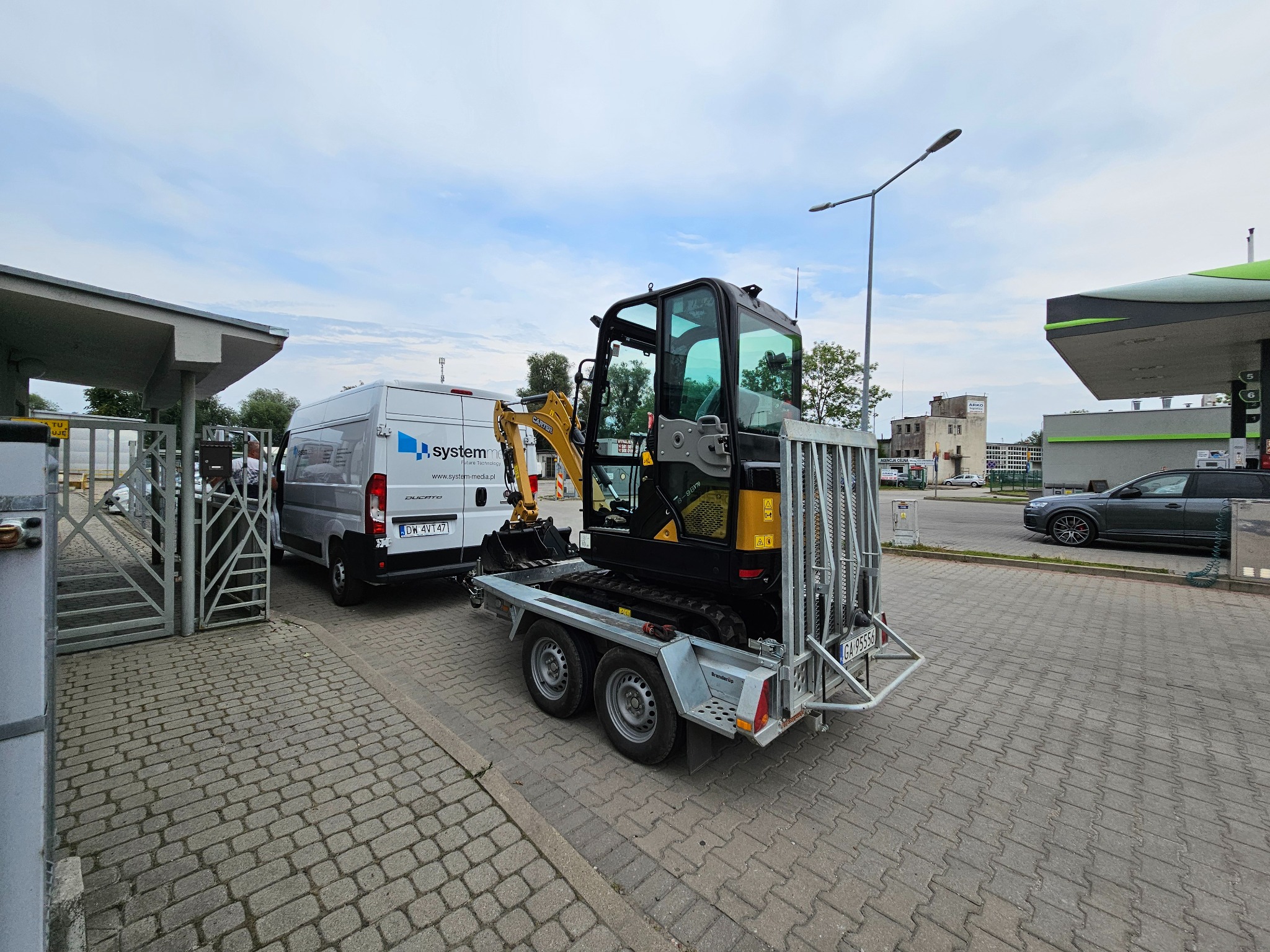 Żółto-czarna mini koparka na szarej przyczepie, zaparkowana obok białego busa SystemME na kostce brukowej. W tle stacja benzynowa i fragment metalowej bramy.