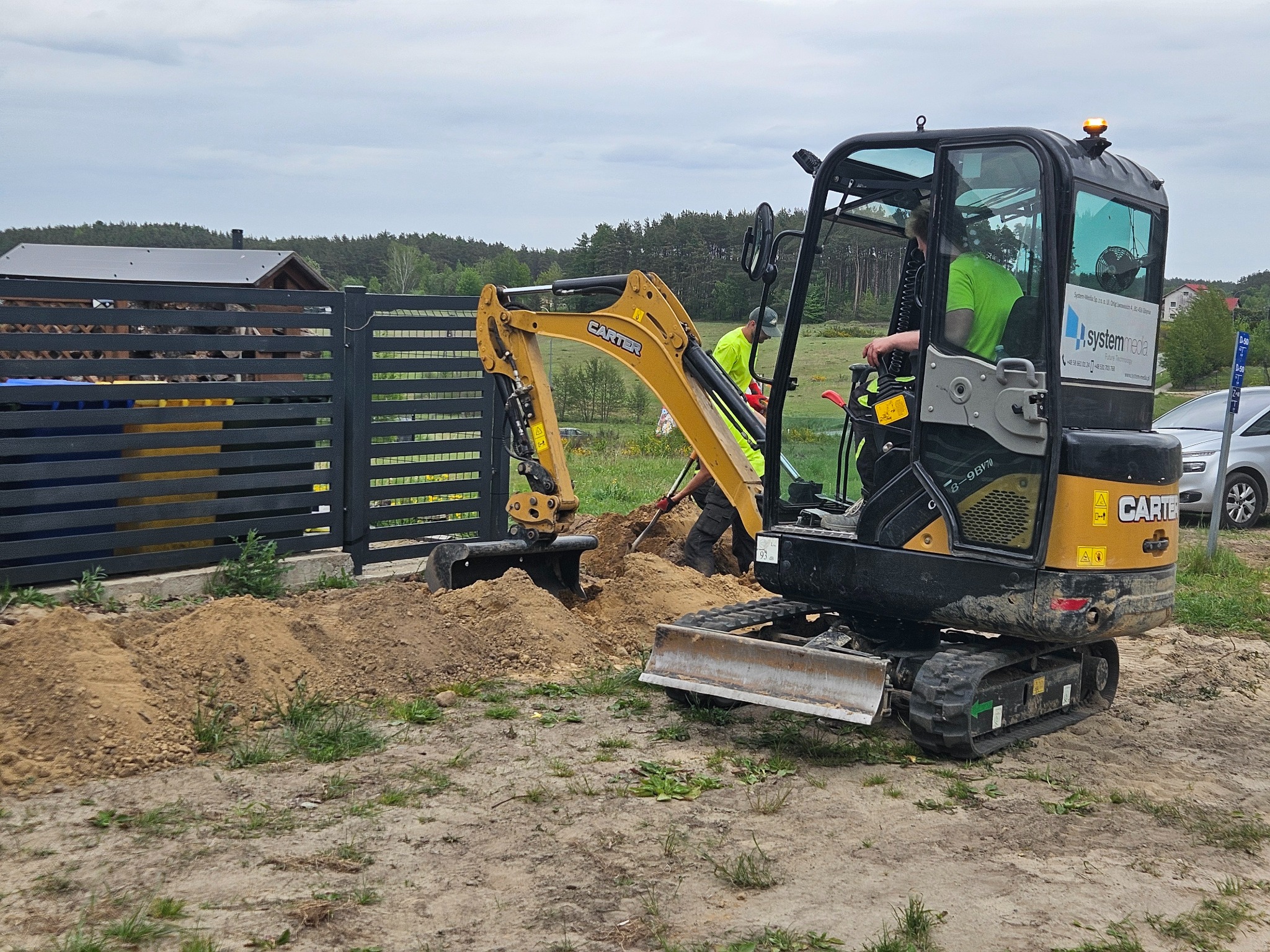 Żółto-czarna mini koparka Carter podczas wykopu pod instalacje przy ogrodzeniu posesji. Widoczny operator w kabinie i pracownik z łopatą. Praca w toku.