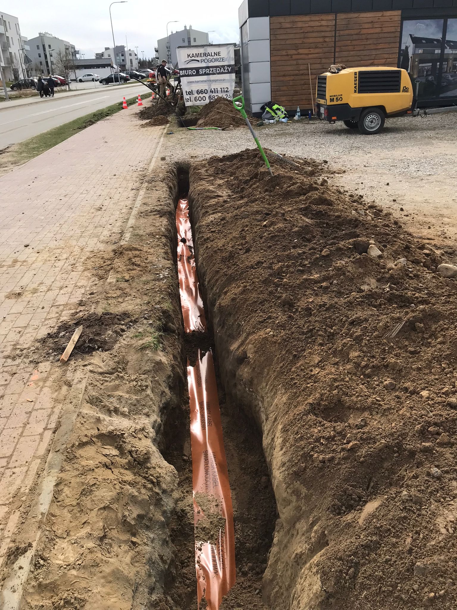 Wykopany rów z pomarańczowymi rurami kablowymi, w tle budynek biura sprzedaży i sprężarka Kaeser M 50. Prace ziemne pod instalacje telekomunikacyjne.
