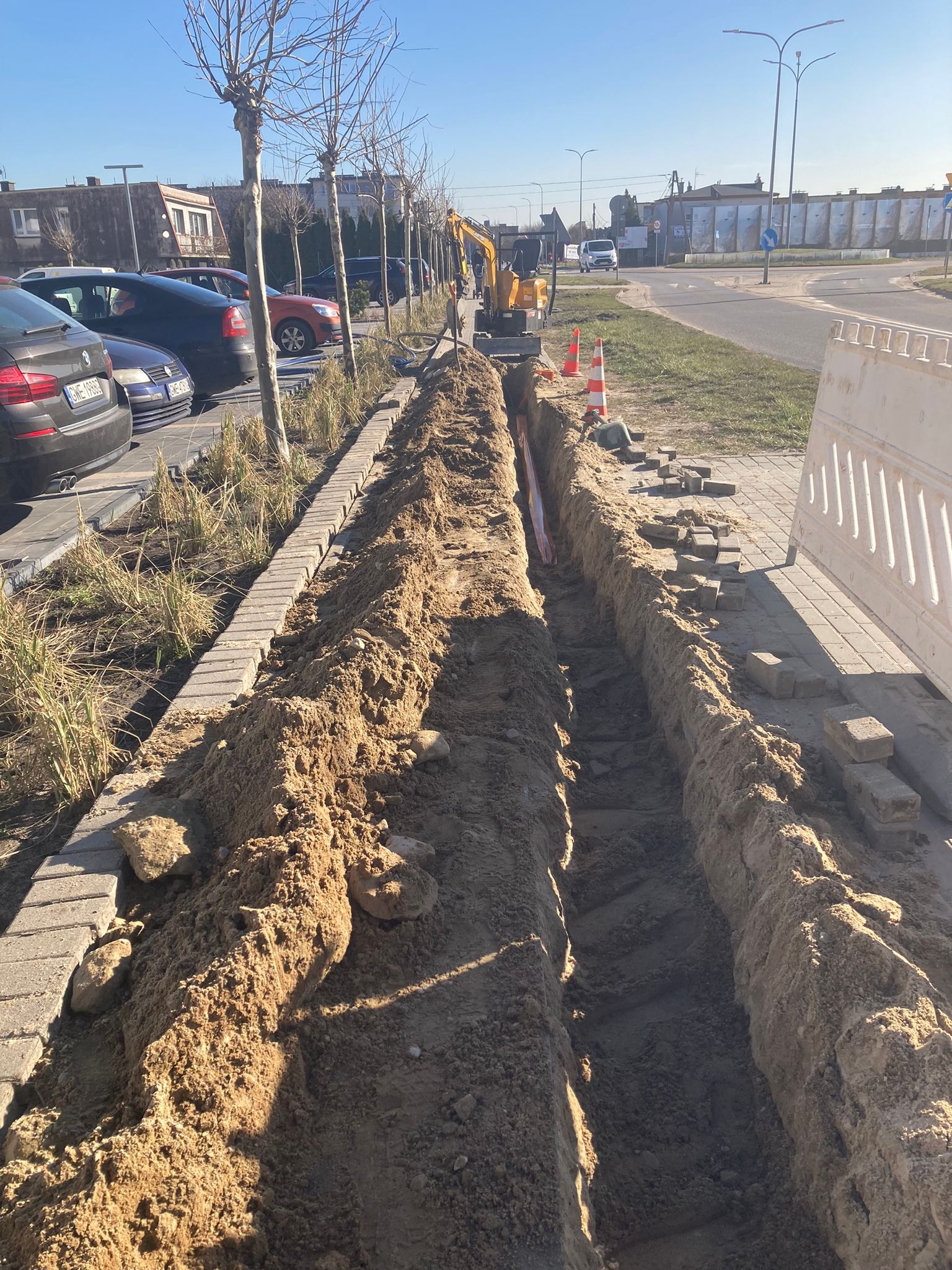 Długi wykop pod instalacje biegnący wzdłuż chodnika, z widocznym mini koparką i pomarańczowymi pachołkami. W tle zaparkowane samochody i budynki.
