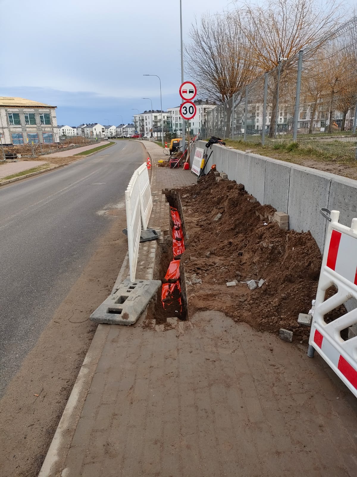 Wykop w chodniku zabezpieczony barierkami, z widocznymi pomarańczowymi osłonami kabli/rur. W tle ulica, budynki i znaki drogowe. Prace ziemne w trakcie realizacji.