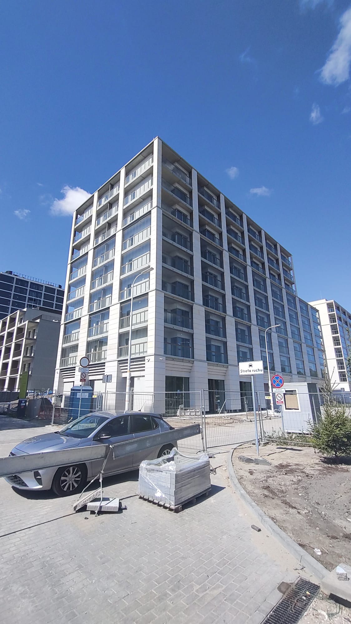 Nowoczesny budynek wielorodzinny w trakcie budowy, widoczny samochód i materiały budowlane. Jasne niebo z delikatnymi obłokami w tle.