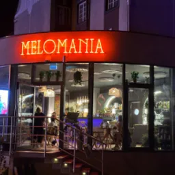 Nocny widok na restaurację Melomania z neonowym napisem, widoczne wnętrze z klientami i barem, schody wejściowe z oświetleniem.