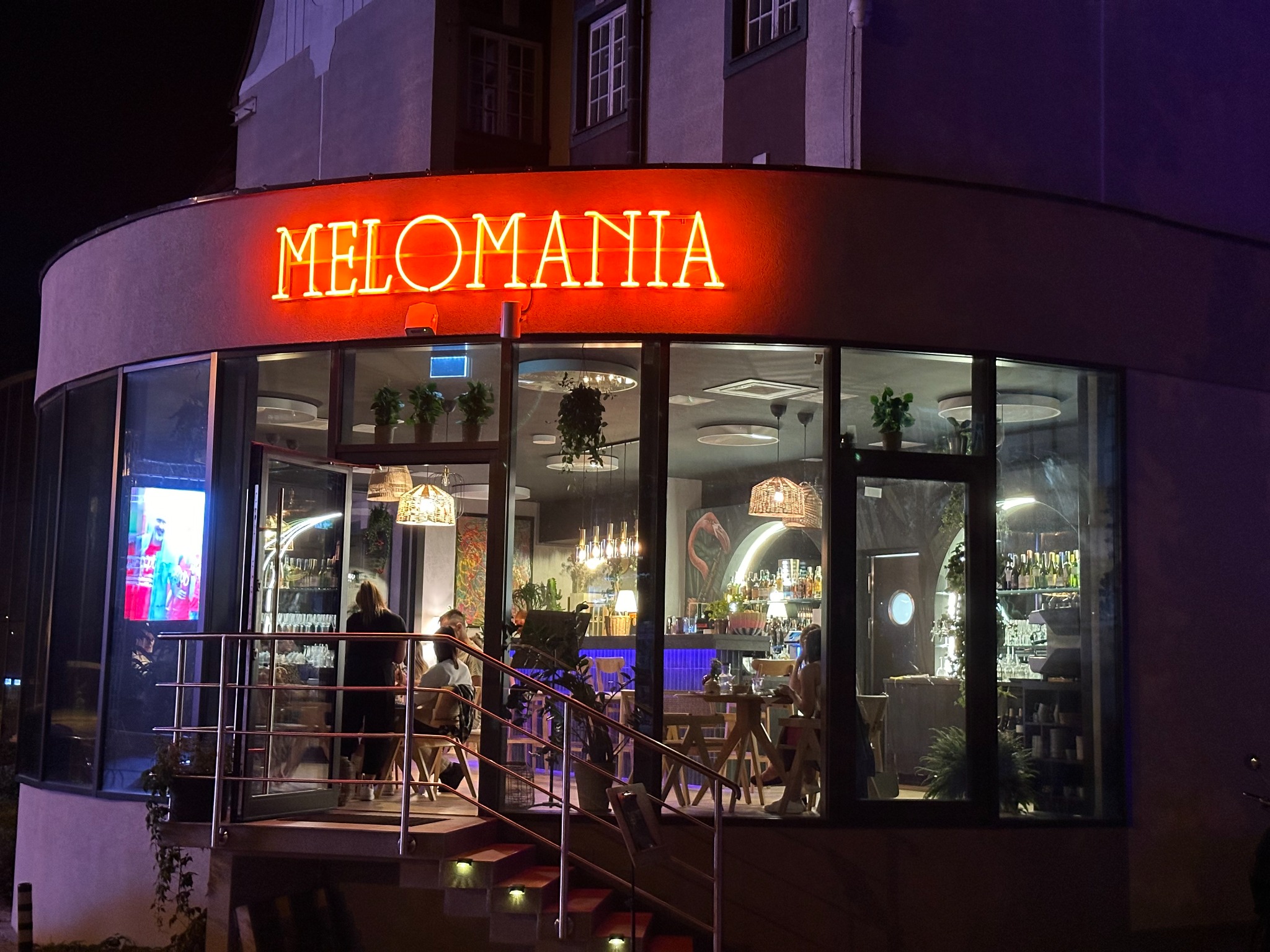 Nocny widok na restaurację Melomania z neonowym napisem, widoczne wnętrze z klientami i barem, schody wejściowe z oświetleniem.