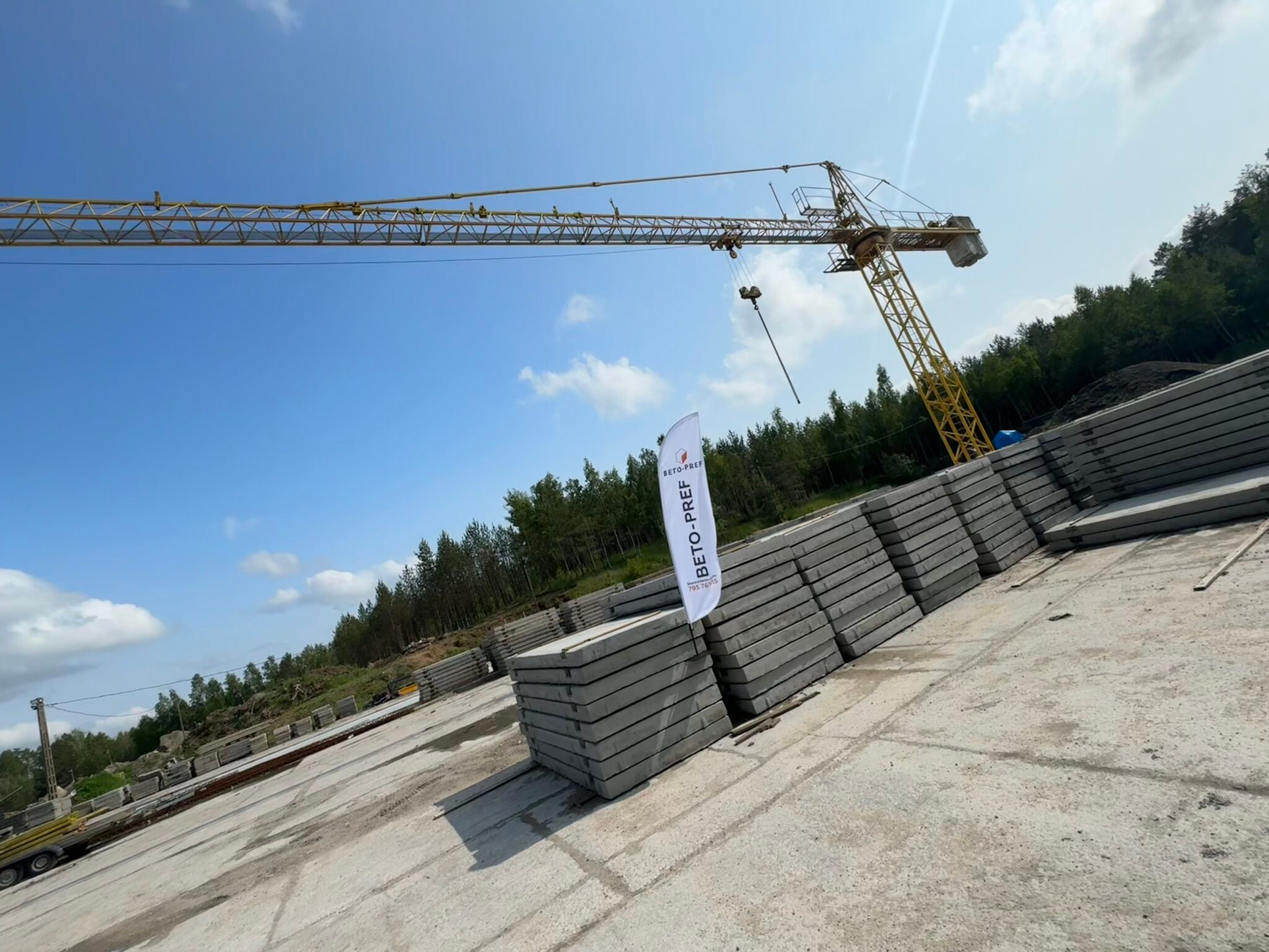 Żuraw budowlany nad stosem szarych płyt betonowych na placu składowym, flaga z logo producenta, zalesione wzgórze w tle, błękitne niebo.