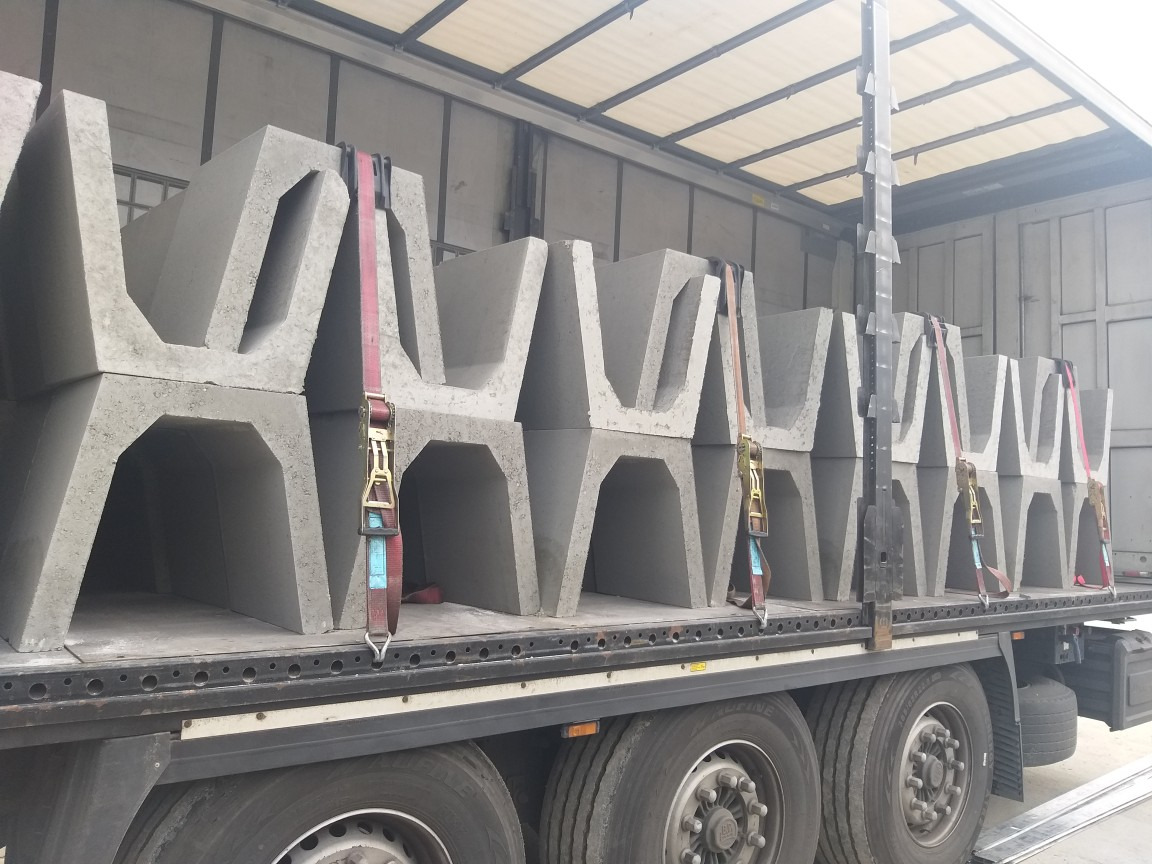 Szare, betonowe prefabrykaty ułożone w rzędach na platformie ciężarówki, zabezpieczone pasami transportowymi. Elementy o charakterystycznym kształcie litery U.