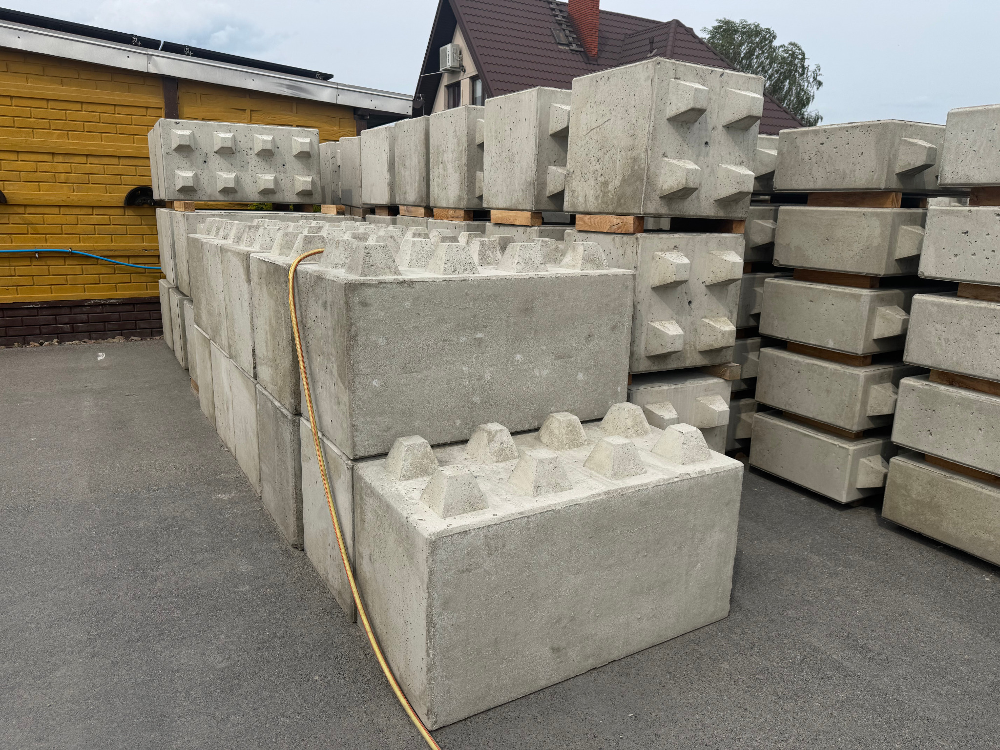 Szare prefabrykaty betonowe ułożone w stosy na zewnątrz, z elementami przypominającymi klocki Lego, na tle budynku z żółtej cegły i domu z brązowym dachem.