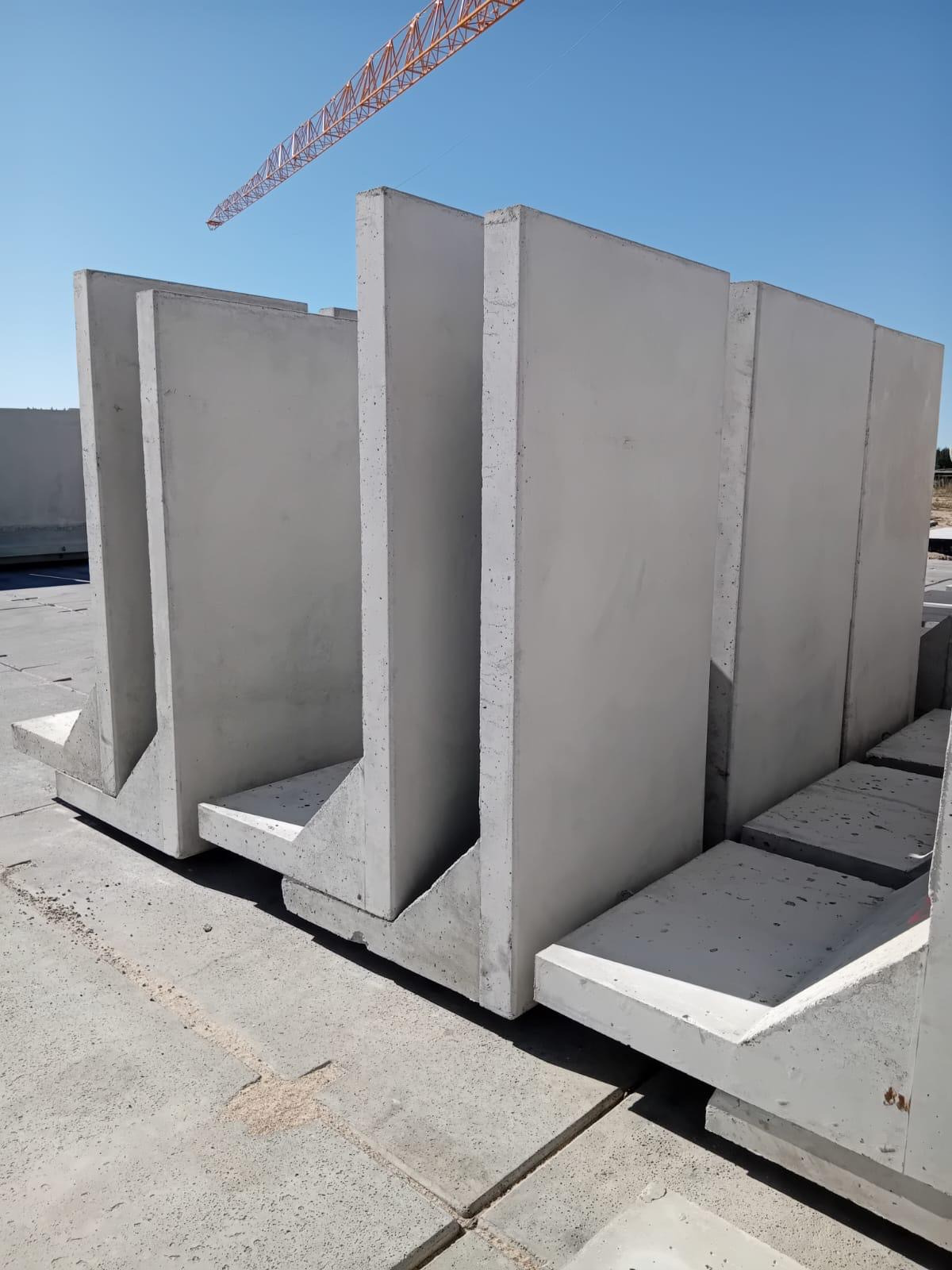 Szare prefabrykaty betonowe w kształcie litery T, ustawione pionowo, widoczne na placu budowy z żurawiem w tle, oświetlone naturalnym światłem słonecznym.