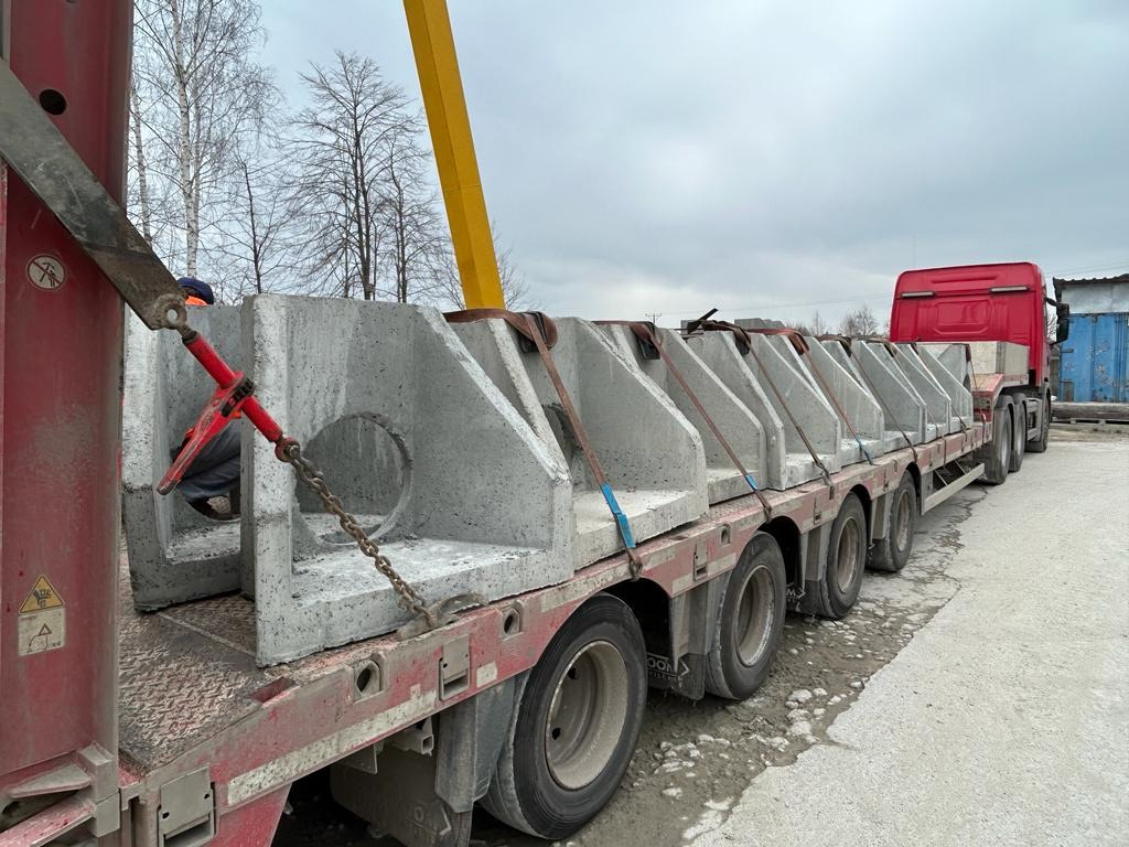 Transport betonowych prefabrykatów na naczepie ciężarówki, widoczne elementy z otworem i ukośnym wzmocnieniem, zabezpieczone łańcuchami, w tle drzewa i pochmurne niebo.