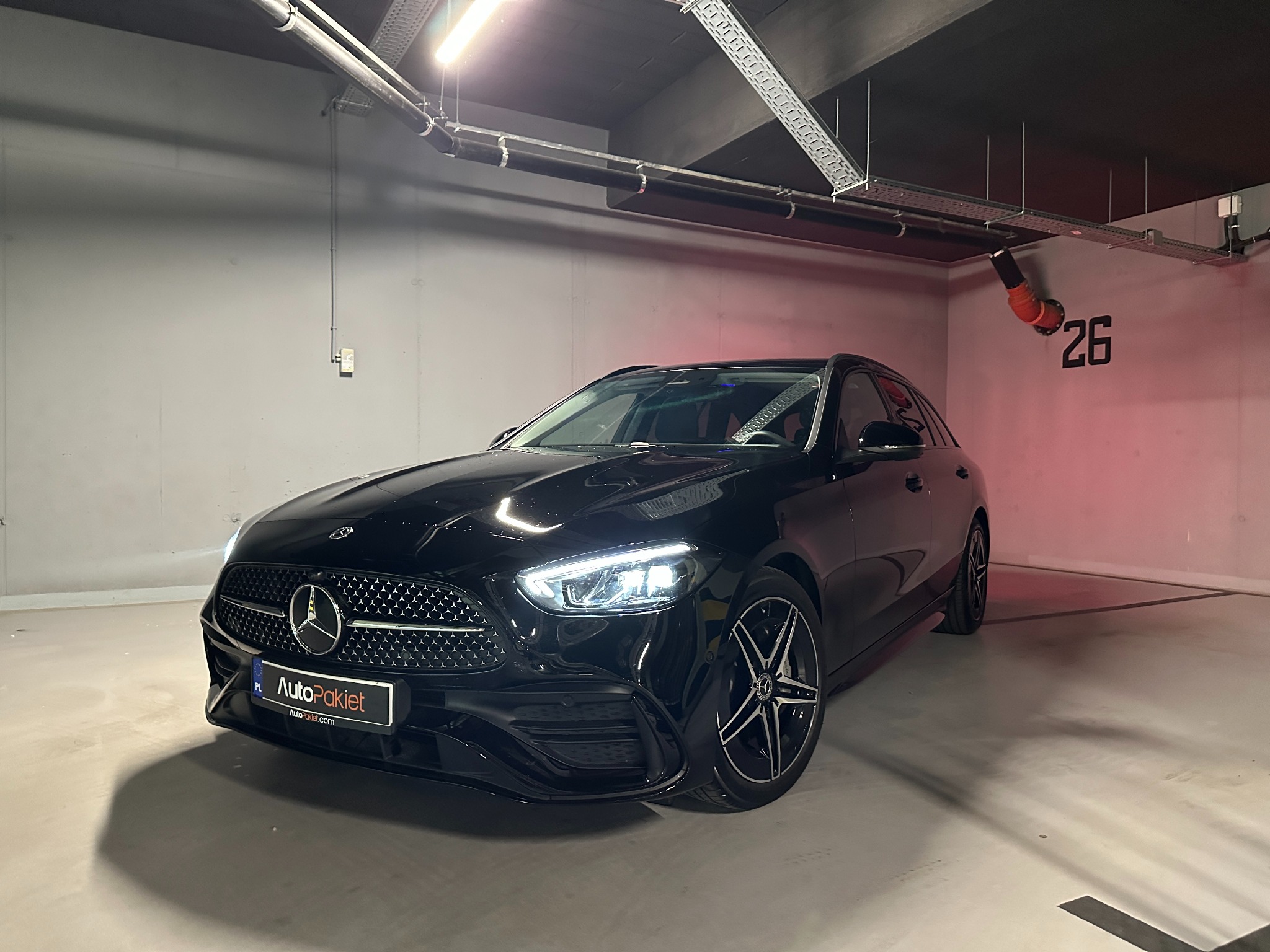 Czarny Mercedes-Benz kombi w garażu podziemnym, widok z przodu, reflektory włączone, oświetlenie sufitowe i numer stanowiska 26 w tle.