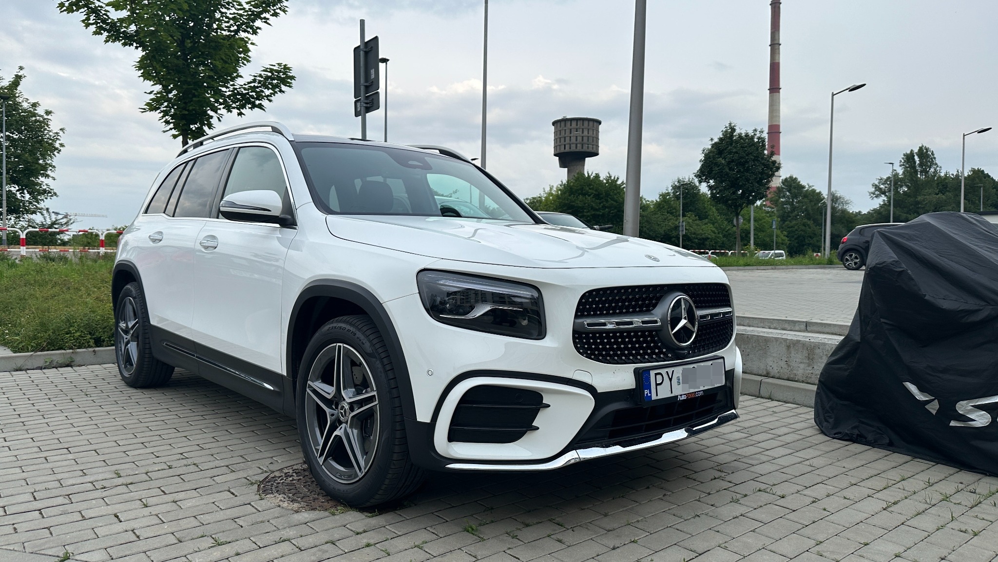 Biały Mercedes-Benz GLB na kostce brukowej, widok z przodu, na tle zieleni i miejskiej infrastruktury. Auto z polską tablicą rejestracyjną.