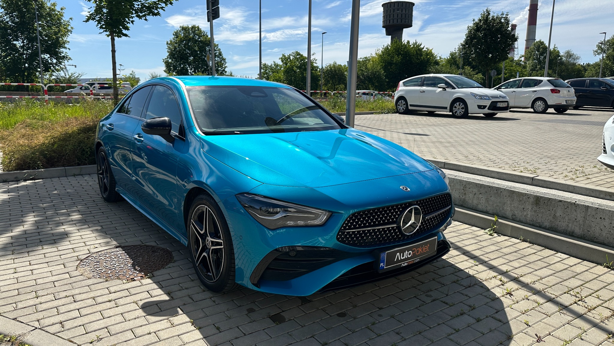 Błękitny Mercedes CLA na parkingu, z widocznym logo 'AutoPakiet' na tablicy rejestracyjnej. W tle inne pojazdy i wieża ciśnień.