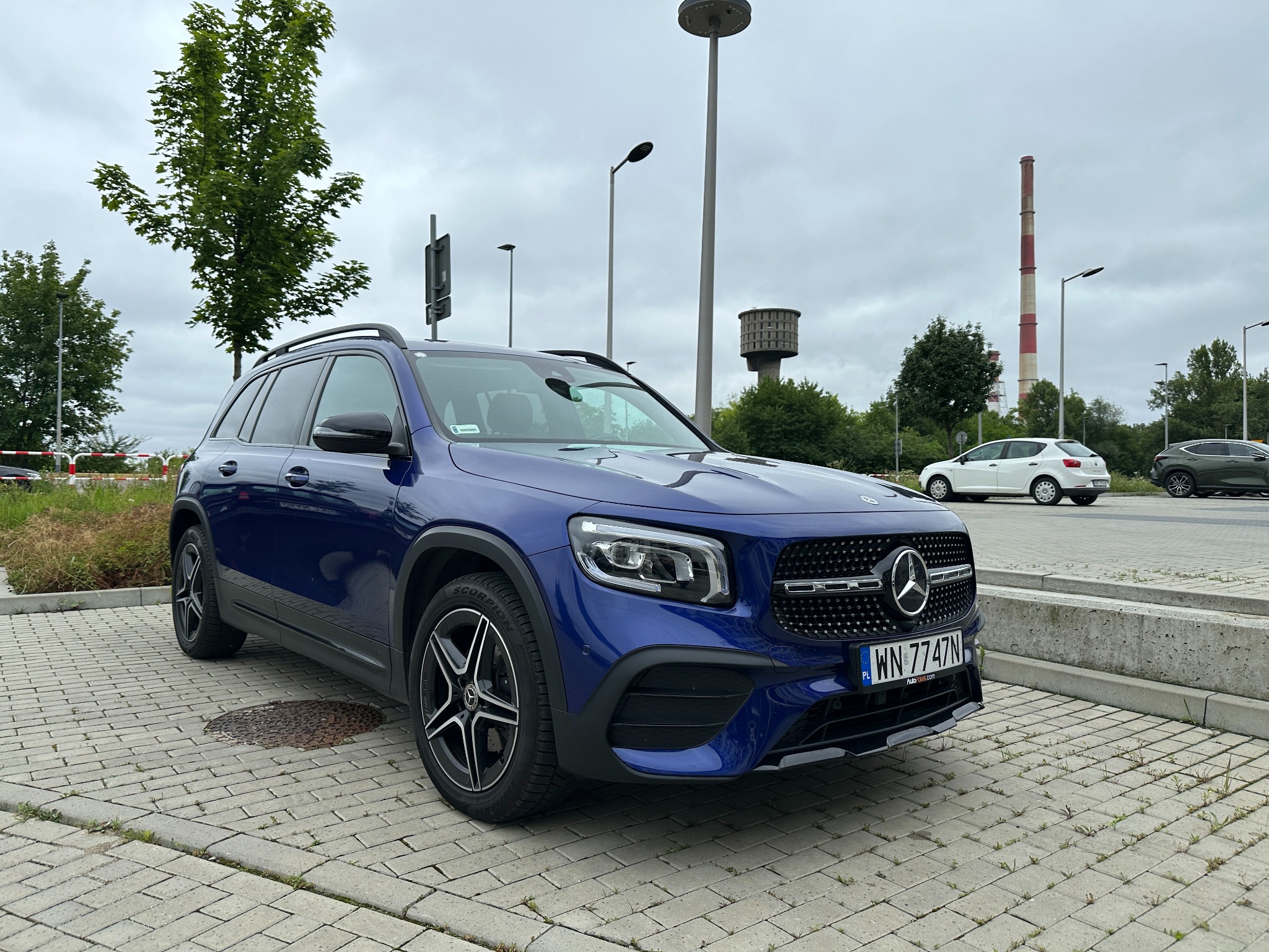 Niebieski Mercedes-Benz GLB na brukowanym parkingu z drzewami, kominem przemysłowym i wieżą ciśnień w tle. Tablica rejestracyjna z przodu.