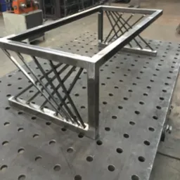 Stalowa konstrukcja ramy stołu z ukośnymi prętami, spawana na perforowanym stole spawalniczym. Widoczne ślady spawania i obróbki.