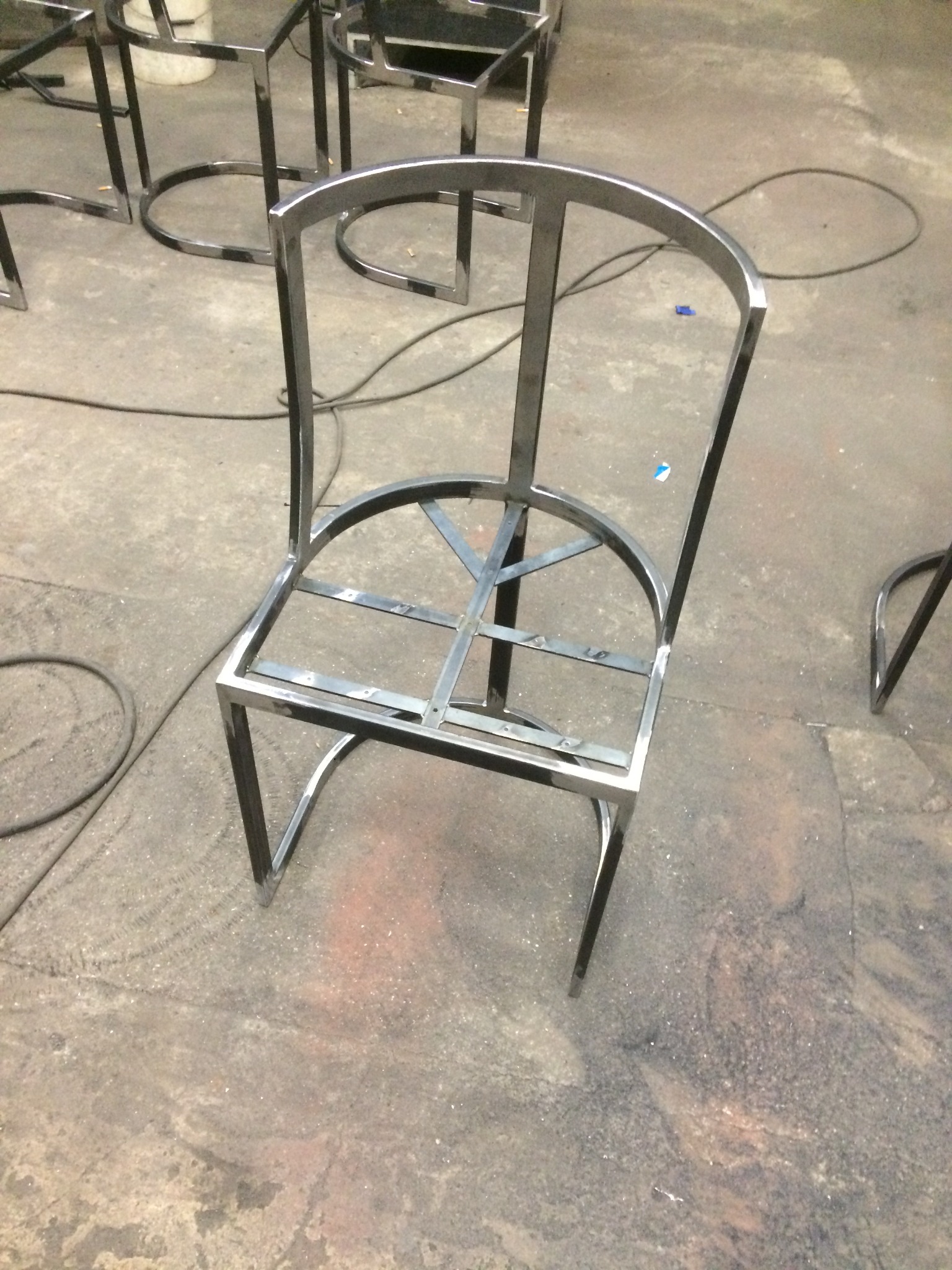 Metalowy szkielet krzesła z rurek, spawany, na szorstkiej posadzce. Kilka podobnych konstrukcji w tle. Widoczne ślady spawania.