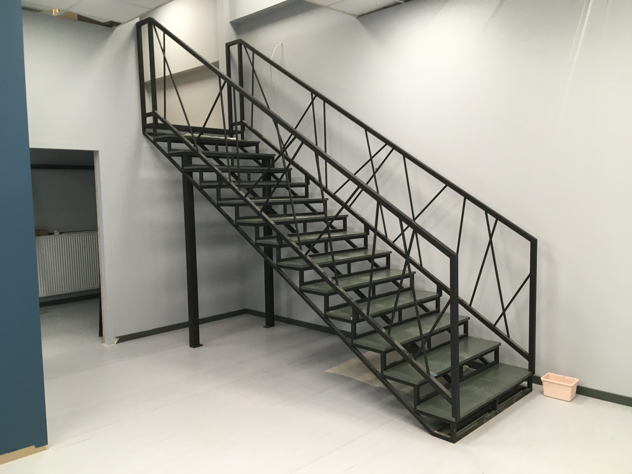 Nowoczesne schody metalowe z czarną, geometryczną balustradą, umieszczone w minimalistycznym wnętrzu z jasnymi ścianami i podłogą. Widoczne detale konstrukcyjne.